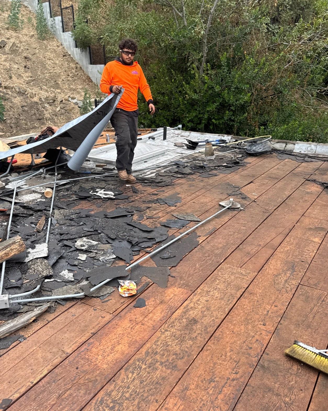torch-down-flat-roof-replacement-insulation-los-angeles-3343 Roofer in orange hoodie peeling black roofing membrane off wooden roof deck with debris scattered around.