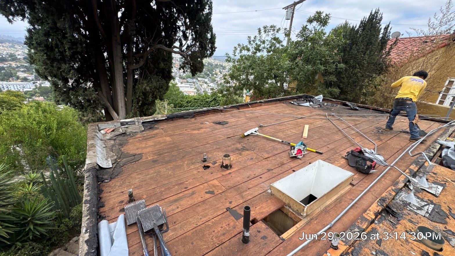 torch-down-flat-roof-replacement-insulation-los-angeles-3345 Roofer on flat roof tear-off exposing wood sheathing, tools and skylight opening visible, hillside neighborhood background.