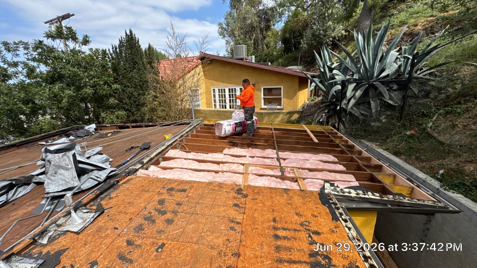 torch-down-flat-roof-replacement-insulation-los-angeles-3346 Roofer placing pink fiberglass insulation batts between wood roof rafters during flat roof renovation, OSB decking visible.