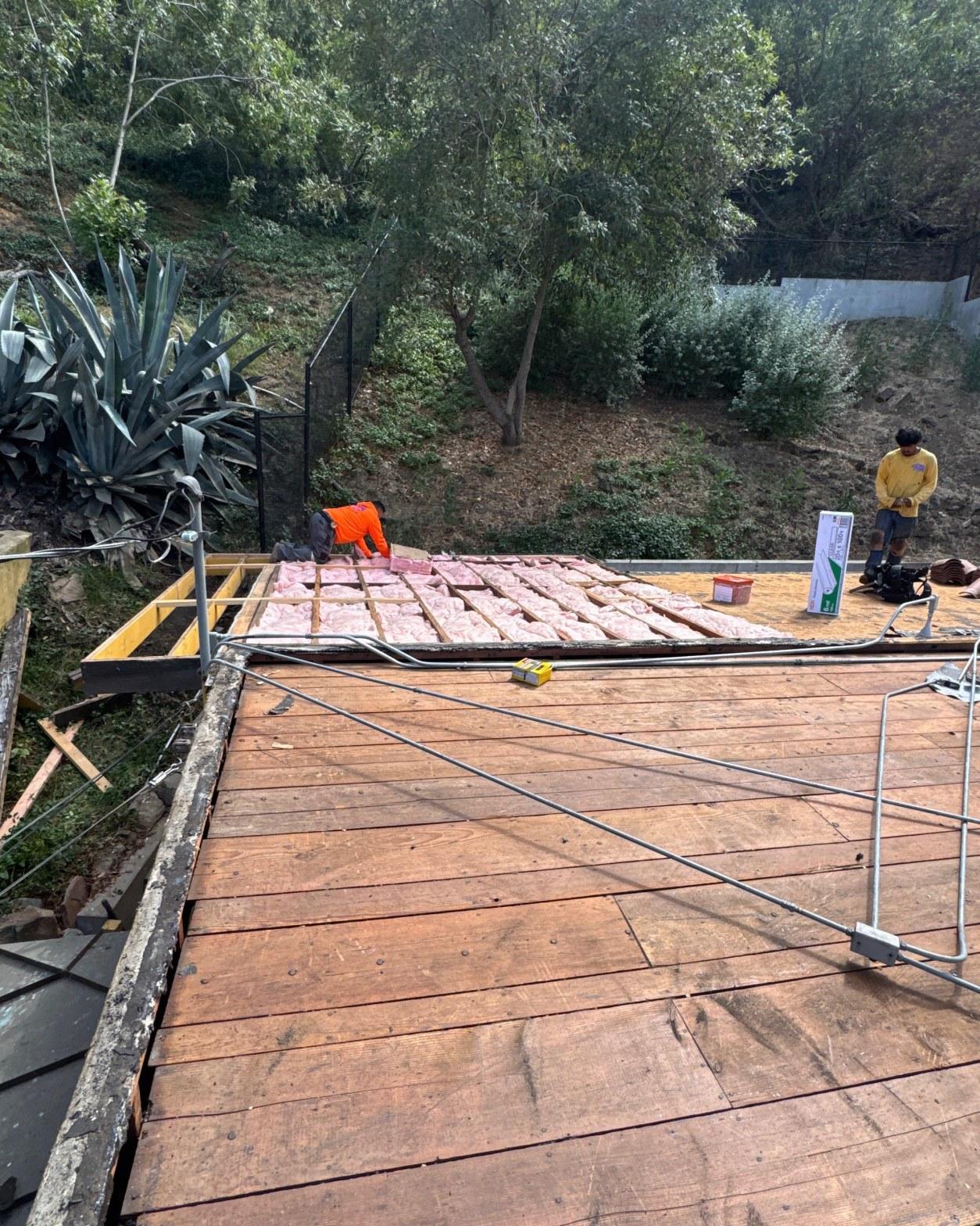torch-down-flat-roof-replacement-insulation-los-angeles-3349 Two roofers placing pink fiberglass insulation batts between wood joists on flat roof deck, green hillside background.