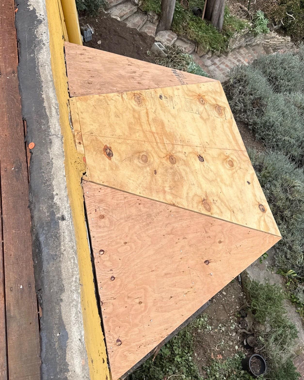torch-down-flat-roof-replacement-insulation-los-angeles-3353 Fresh plywood panels screwed onto sloped roof deck beside yellow steel beam, garden visible below.
