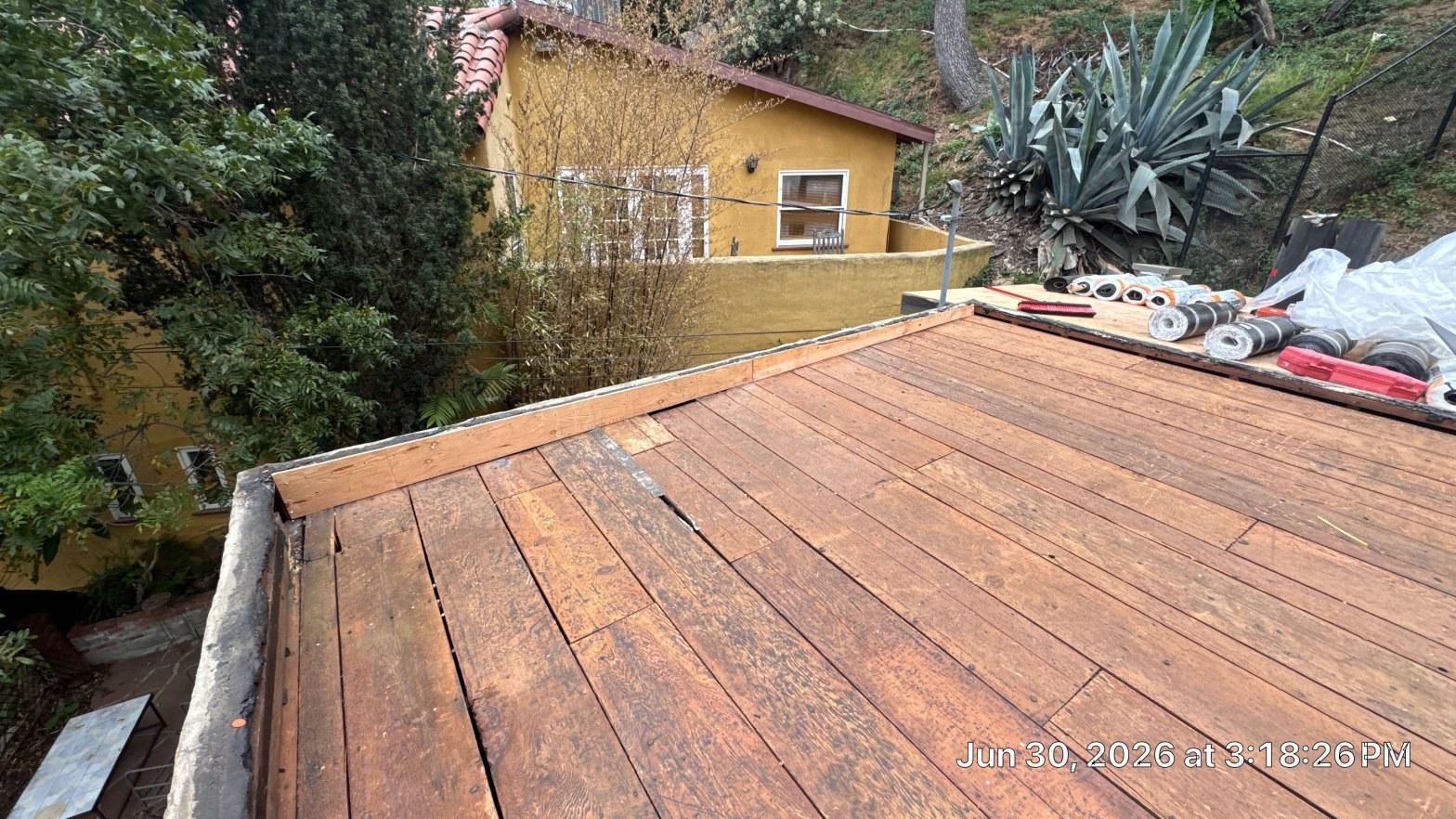 torch-down-flat-roof-replacement-insulation-los-angeles-3354 Bare wood plank flat roof decking with rolled roofing materials on edge, yellow stucco building in background.
