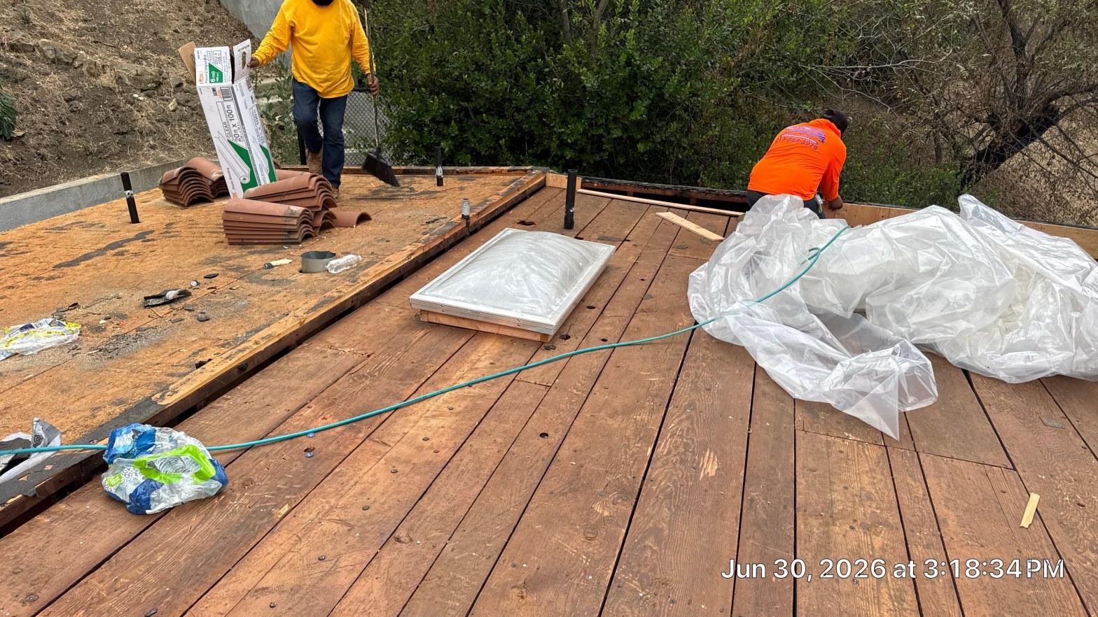 torch-down-flat-roof-replacement-insulation-los-angeles-3355 Two workers on wood roof deck with skylight, rolled underlayment, clay tiles, and plastic sheeting during roofing project.