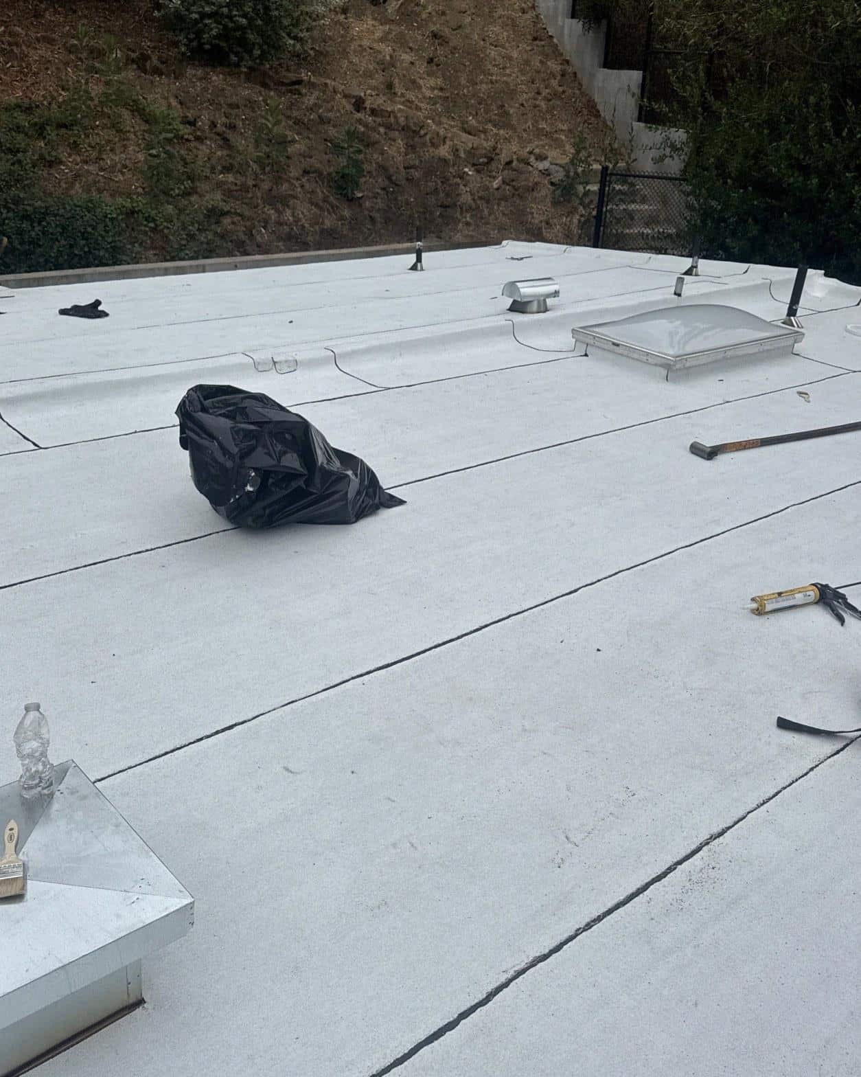 torch-down-flat-roof-replacement-insulation-los-angeles-3358 White TPO flat roof membrane with skylight, roof vents, caulk gun, pry bar, and black trash bag on residential building.