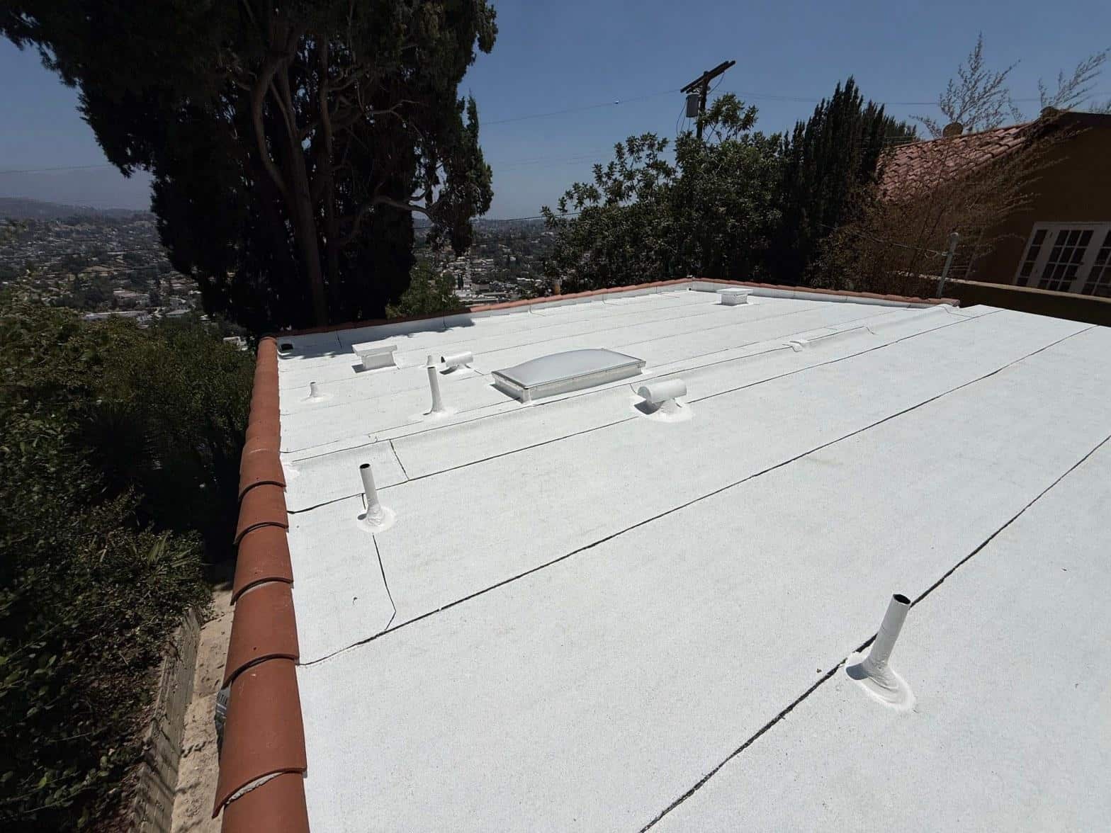 torch-down-flat-roof-replacement-insulation-los-angeles-3361 Newly coated white flat roof with pipe boots, skylight, and terracotta tile edge on Los Angeles hillside home.