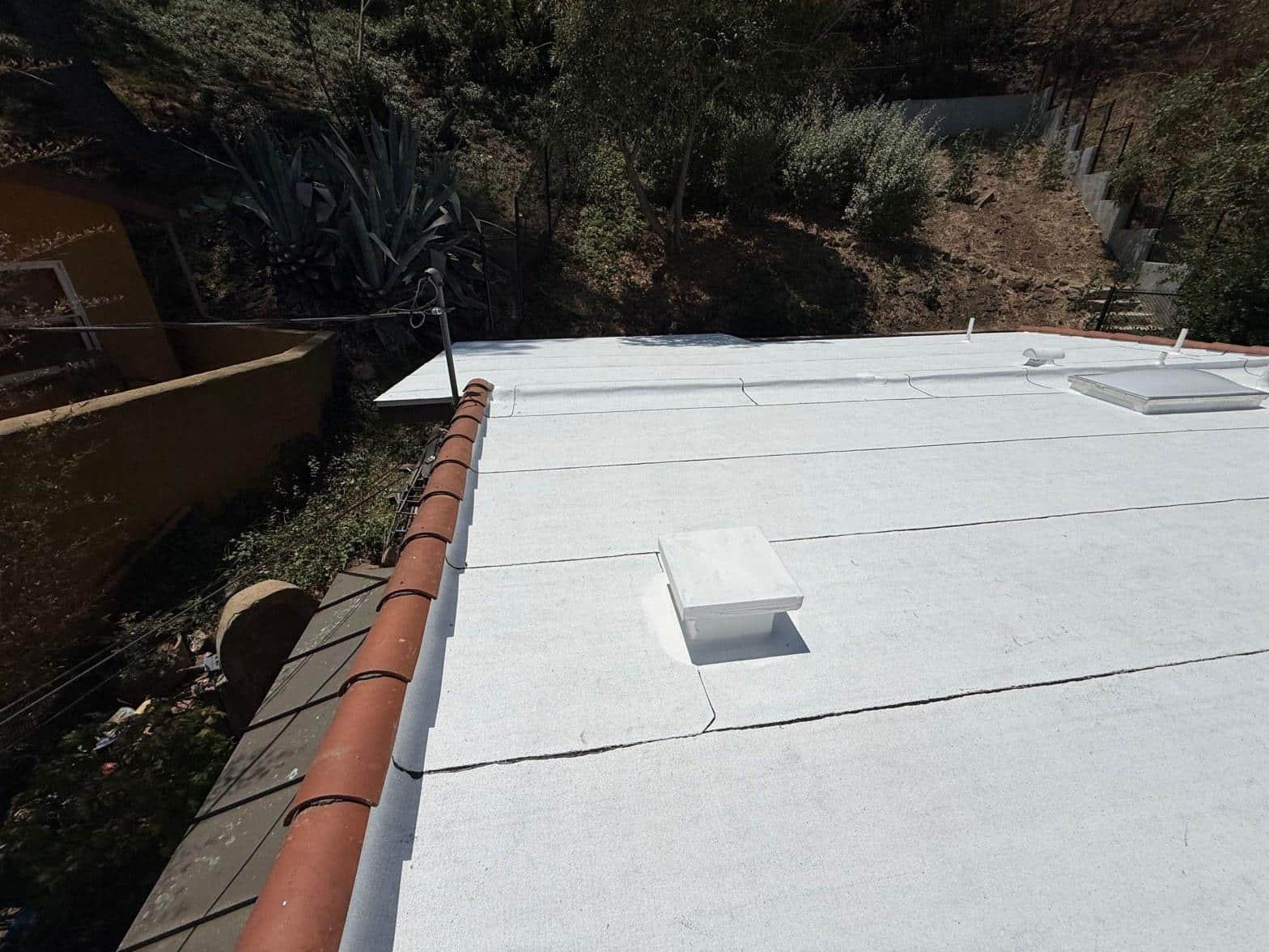torch-down-flat-roof-replacement-insulation-los-angeles-3362 White spray foam flat roof with two skylights, terracotta parapet edge, hillside vegetation in background.