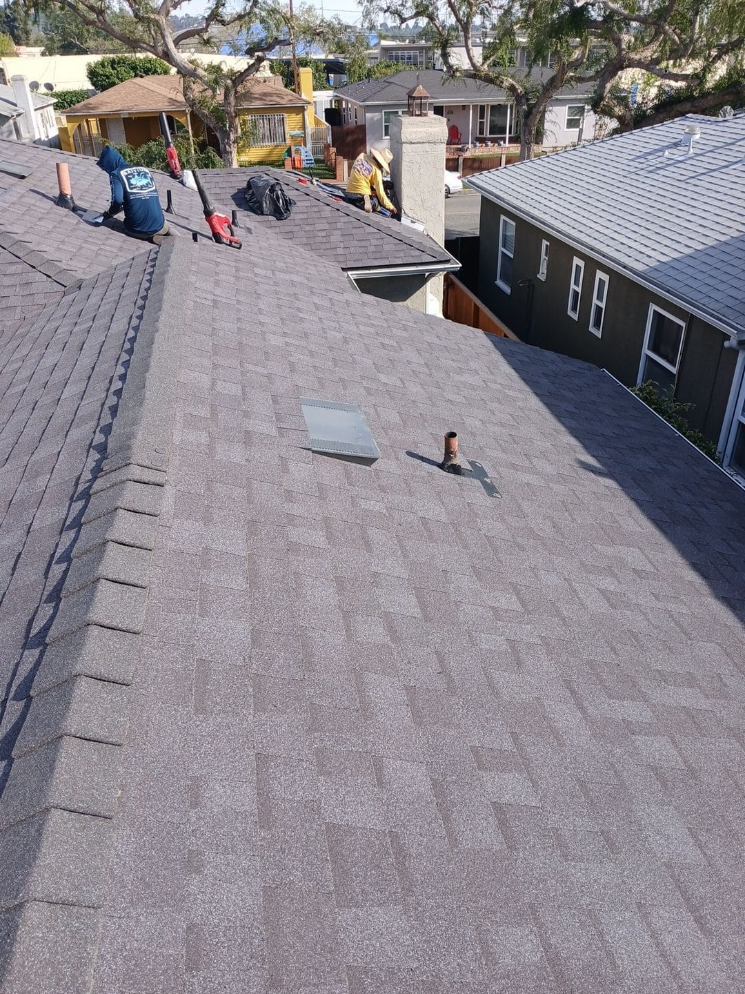 Crew installing gray asphalt shingles on pitched roof with chimney and vent pipe — Roof Replacement Inc