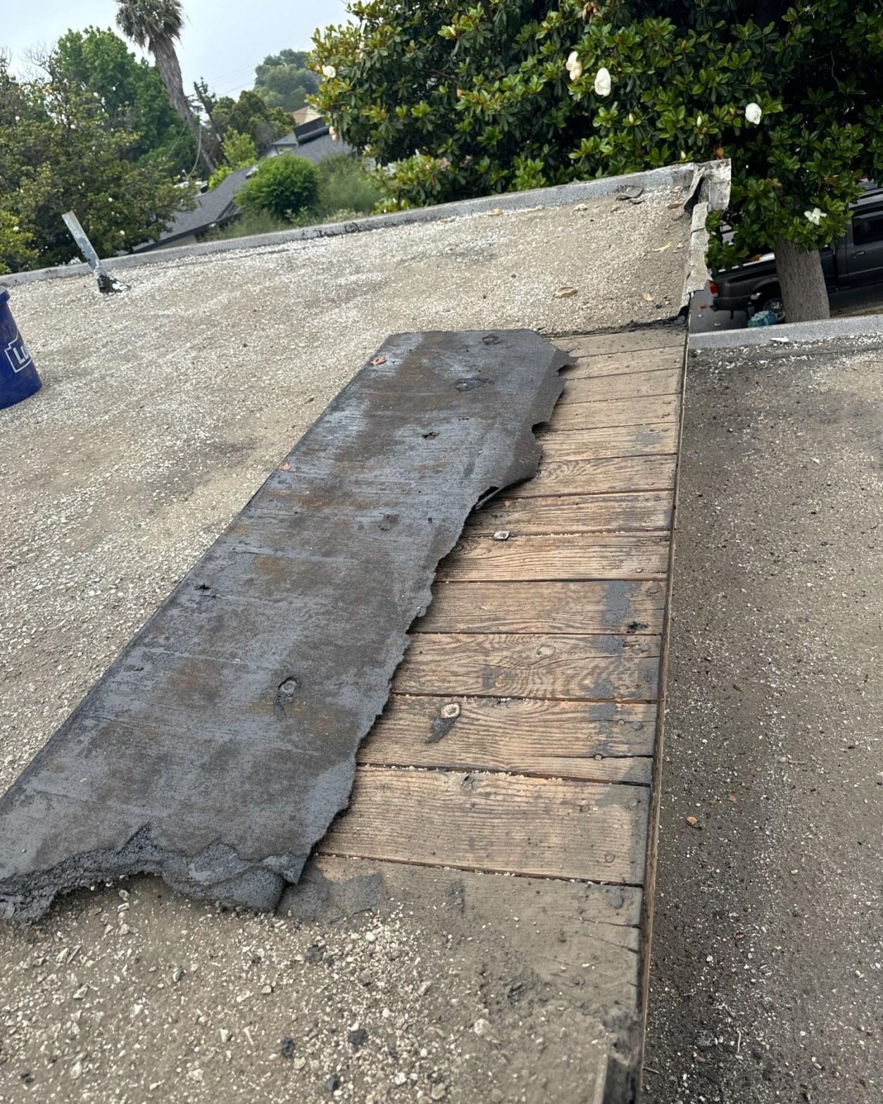 Old gravel-ballasted flat roof membrane peeled back revealing weathered wood plank decking during tear-off