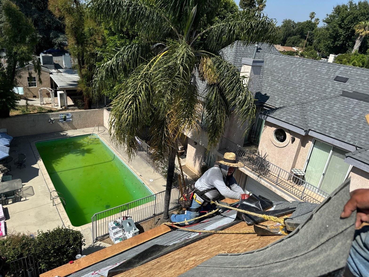 Roofer installing architectural shingles on a lower roof section overlooking a backyard pool during a multi-unit reroof