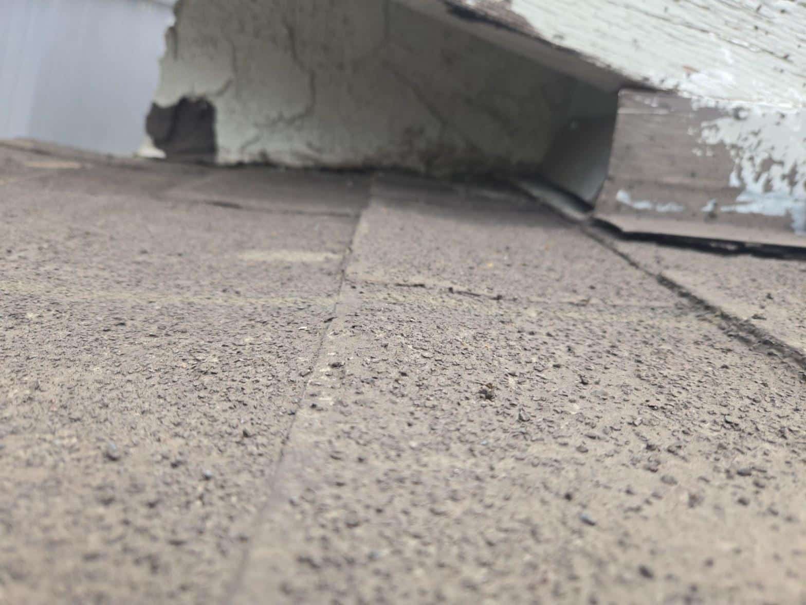 Close-up of heavily granule-depleted asphalt shingles near a wall flashing showing exposed asphalt mat and tab cracking