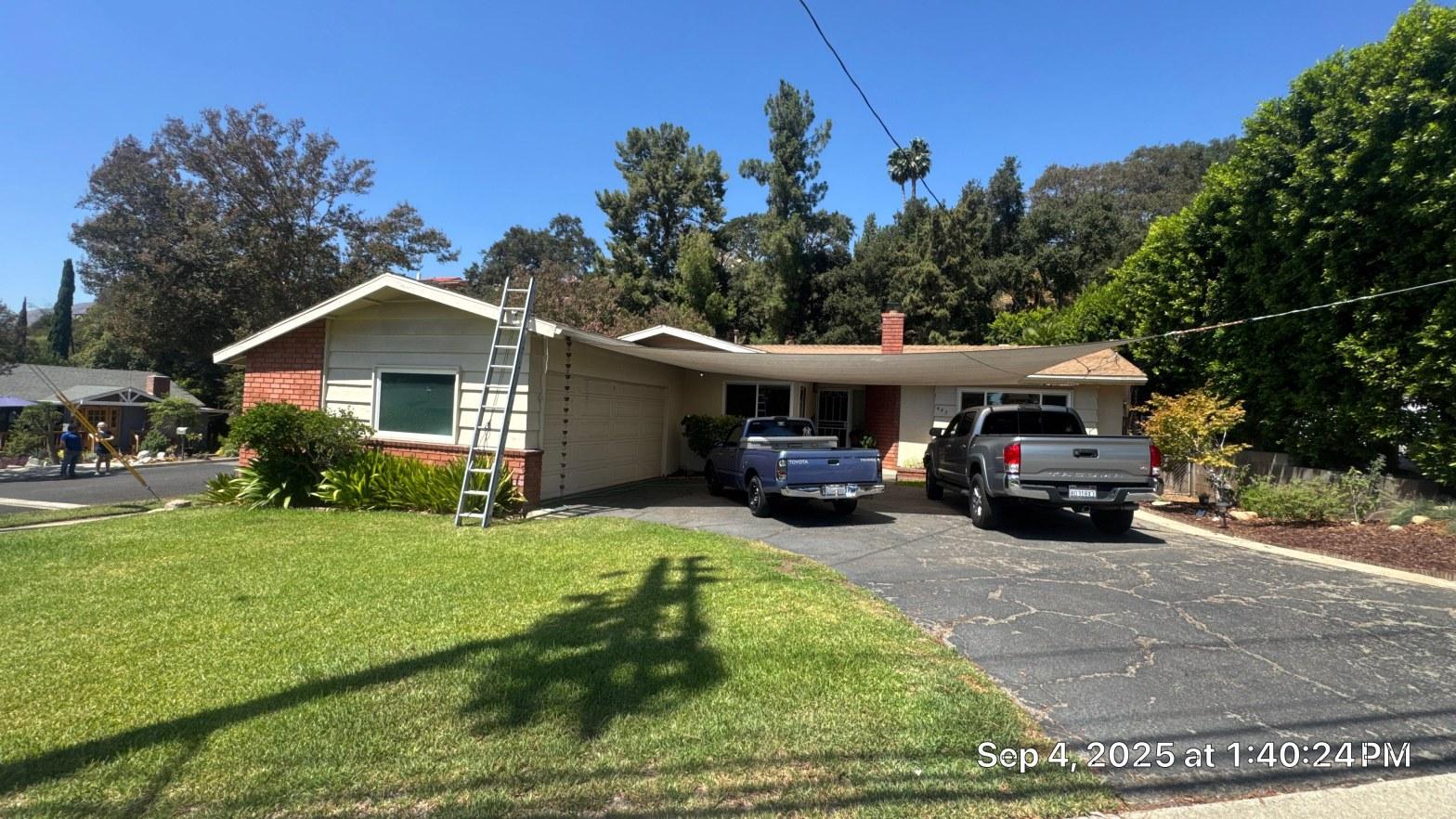 shingle-reroof-monrovia-3386 Extension ladder against single-story brick and stucco ranch house, two Toyota pickup trucks in driveway, sunny day.
