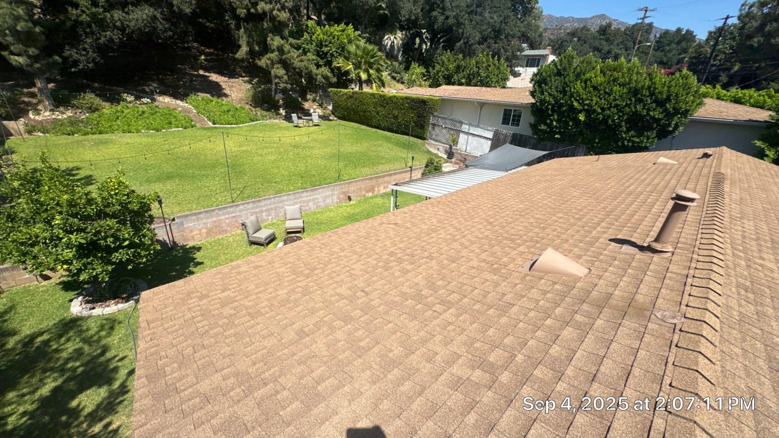 shingle-reroof-monrovia-3389 Newly installed tan asphalt shingle roof with plumbing pipe boots, backyard and hillside visible below.