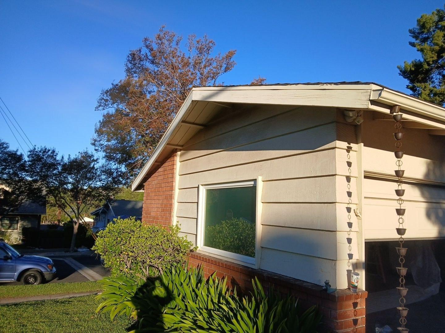 shingle-reroof-monrovia-3394 Decorative copper rain chain downspouts on corner of beige lap siding house with brick foundation and large window.