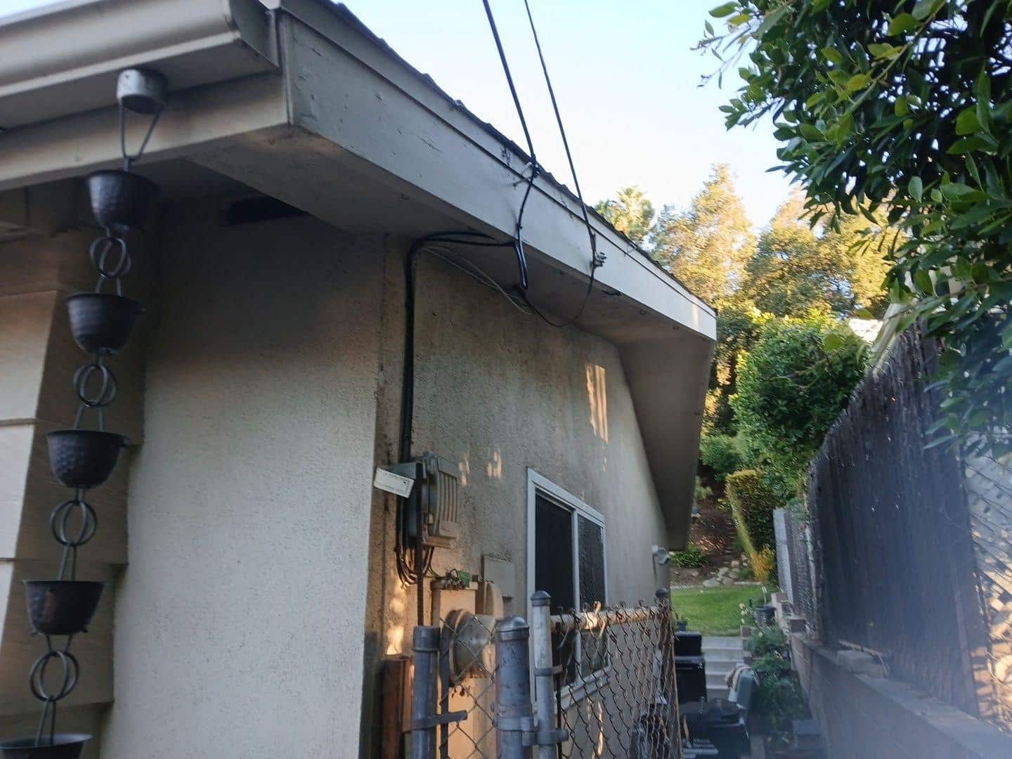 shingle-reroof-monrovia-3396 Decorative copper rain chain attached to gutter on corner of stucco house with chain-link fence and garden.