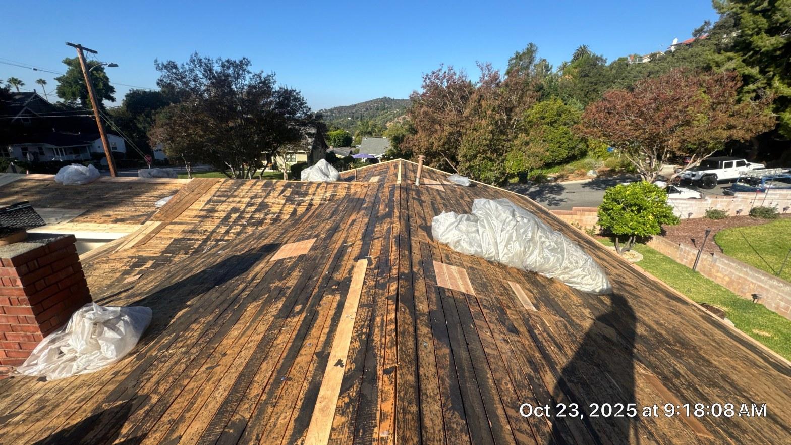 shingle-reroof-monrovia-3400 Stripped roof deck showing aged wood planks, patch boards, and debris bags on residential home, Oct 2025.