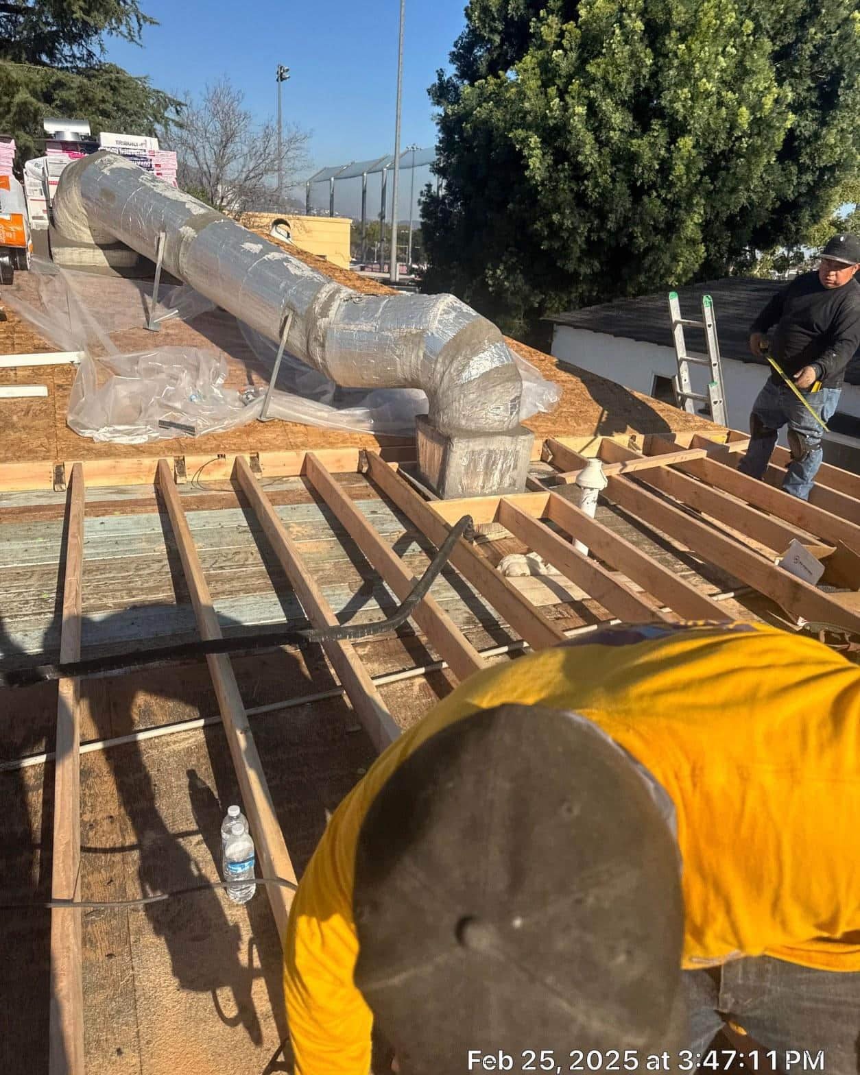 Exposed rafter framing and foil-wrapped duct at ridgeline during reroof with crew measuring near chimney