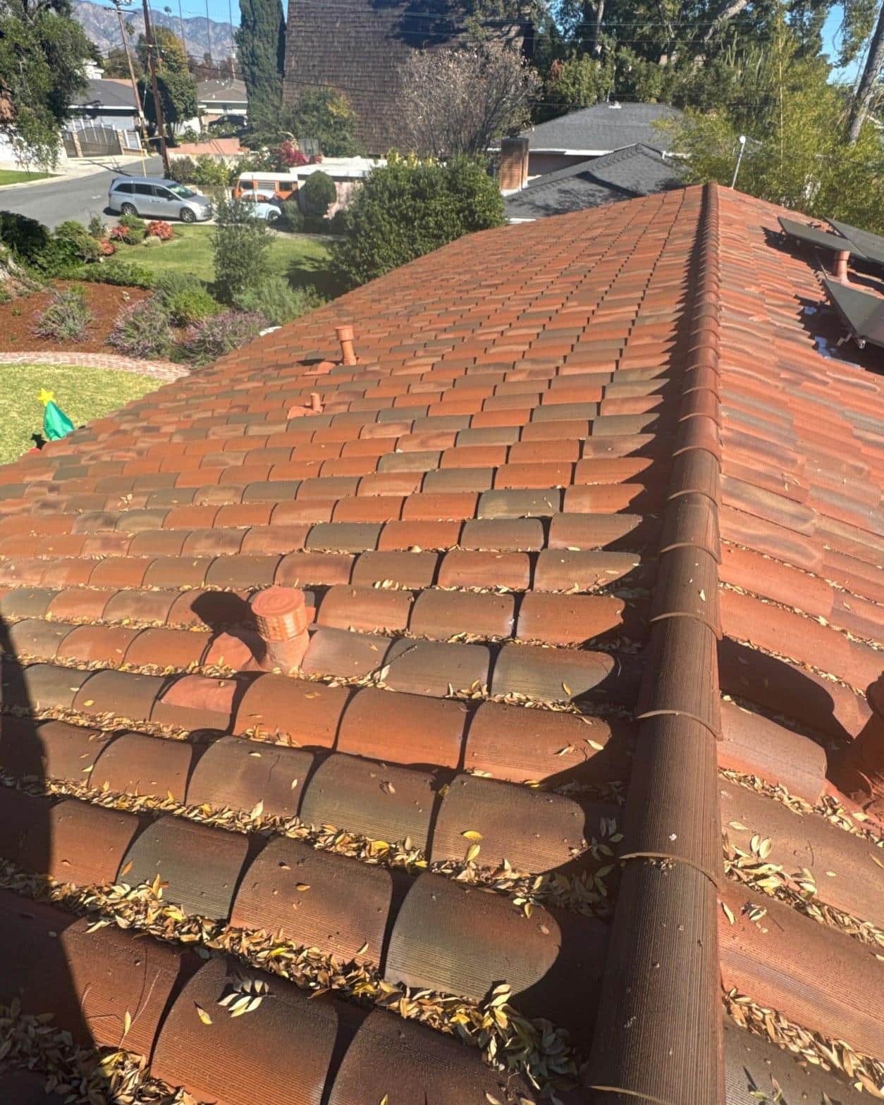 tile-roof-repair-vent-flashing-san-gabriel-3419 Red clay tile roof with dry leaves accumulated in rows and two terracotta plumbing vents, suburban neighborhood background.