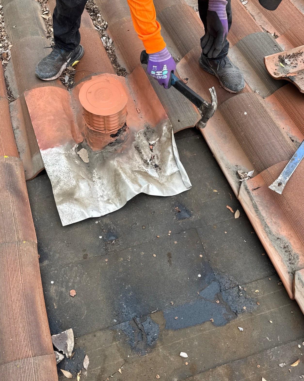 tile-roof-repair-vent-flashing-san-gabriel-3425 Roofers using hammer to lift clay roof tiles around lead flashing and orange soil vent pipe on exposed felt underlayment.