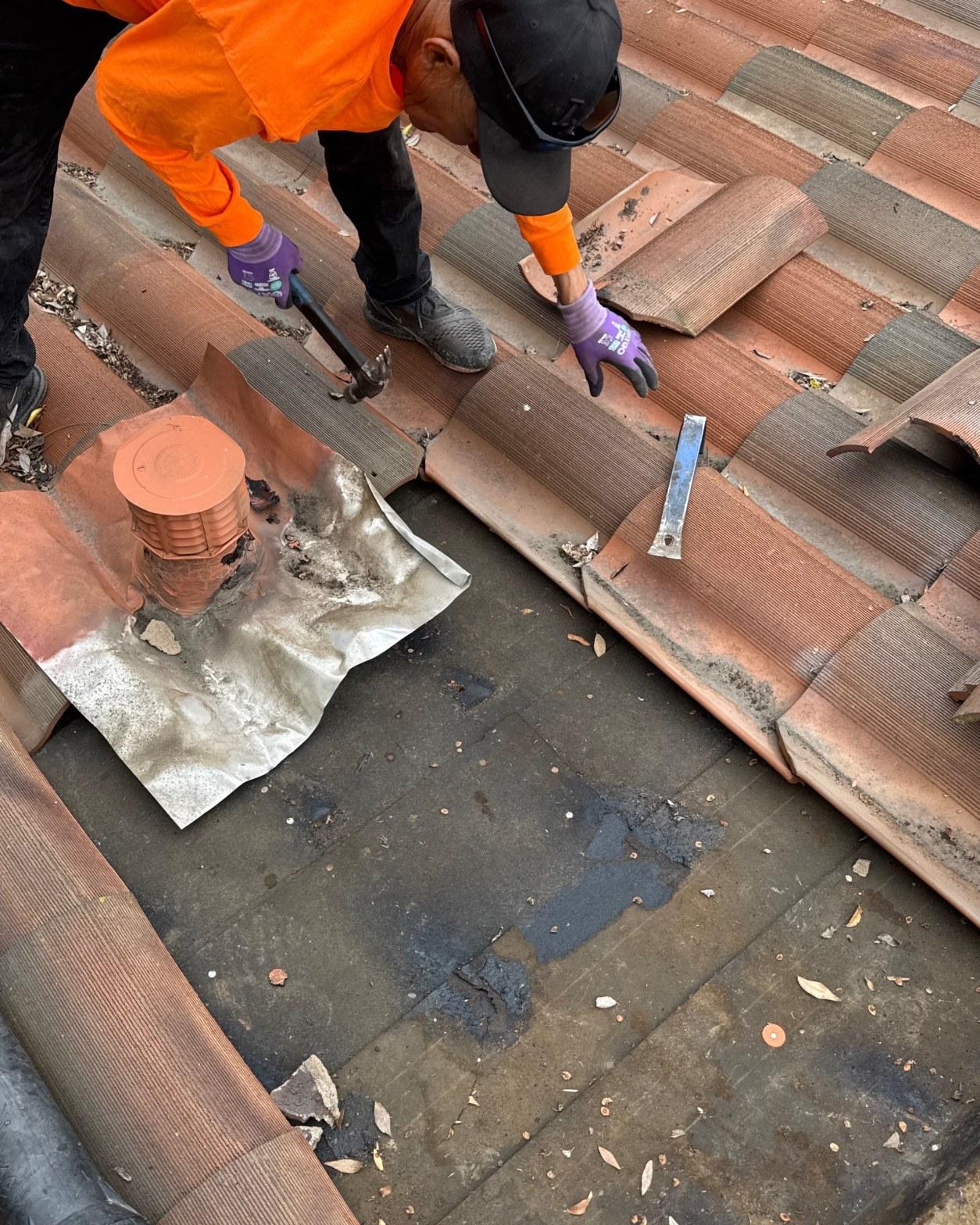 tile-roof-repair-vent-flashing-san-gabriel-3426 Worker in orange high-vis removing terracotta roof tiles, exposing felt underlayment and metal flashing around roof vent.